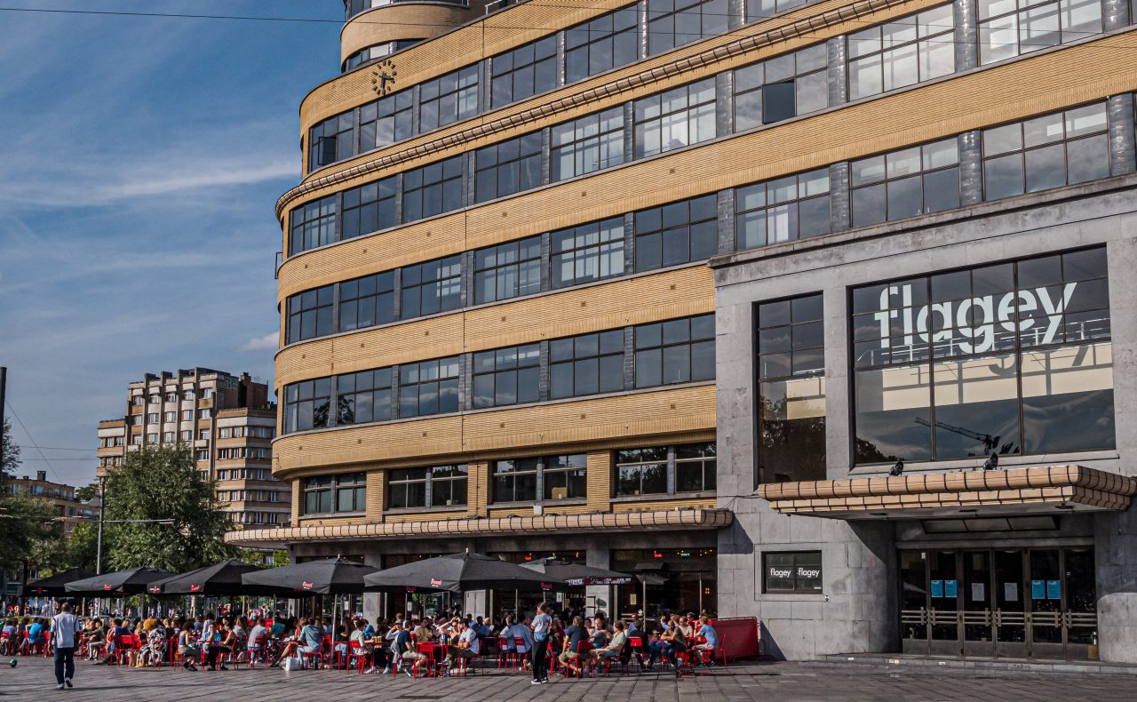 The Flagey building shining in the sun, in the foreground people on the terrace of Café Belga under black parasols